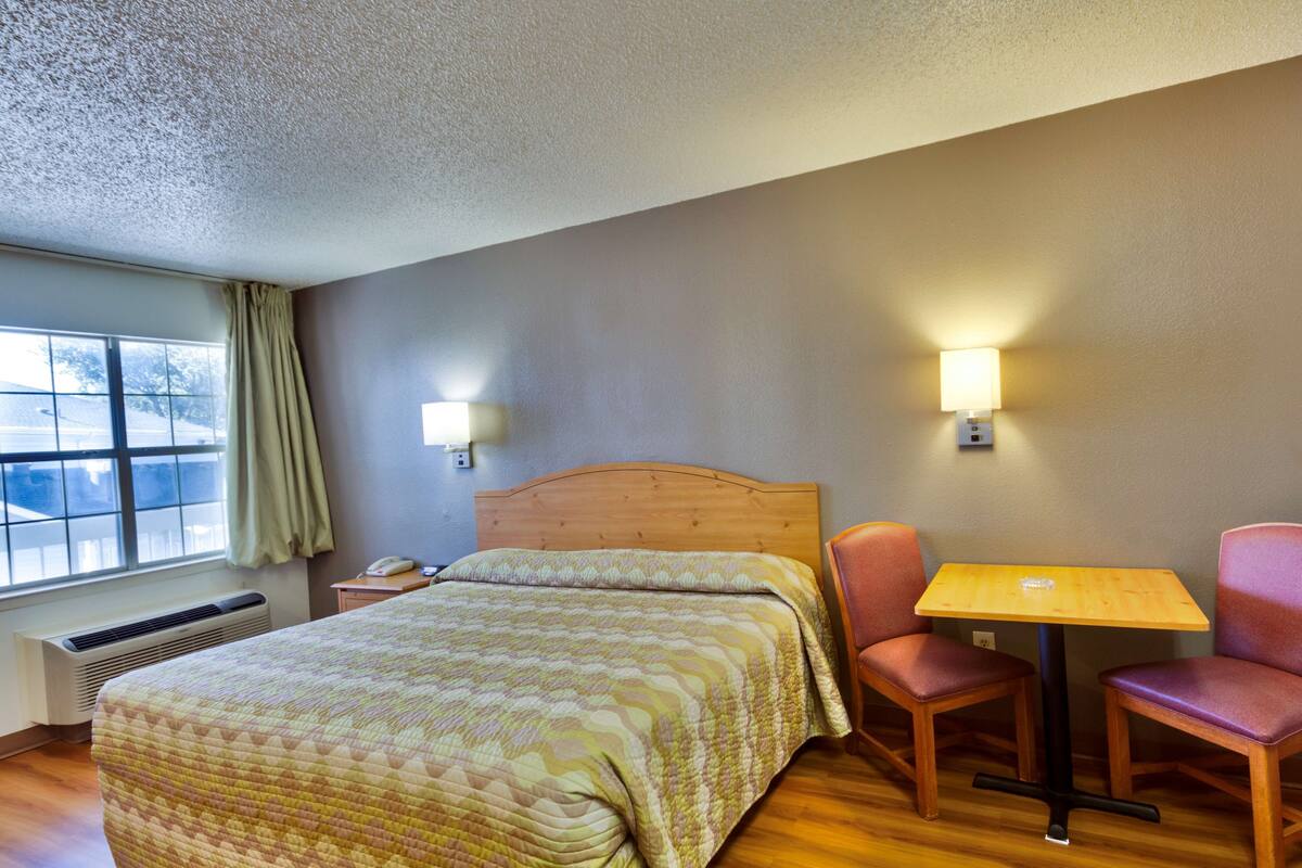 A hotel room with a bed, a table, two chairs, a window with curtains, and wall-mounted lamps.
