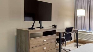 A hotel room with a flat-screen TV mounted on a wooden dresser, a desk with a chair, and a large window with curtains. - Suburban Studios Raleigh - Crabtree Valley (Raleigh)
