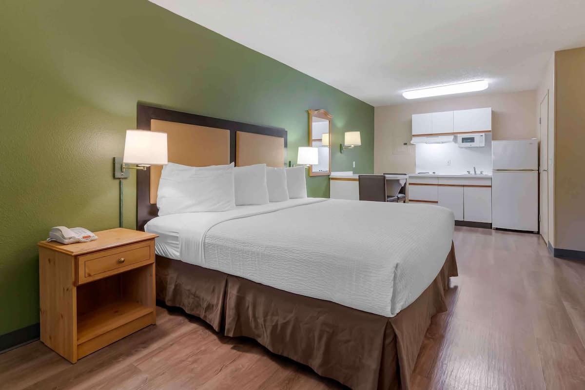 Ein Hotelzimmer mit Bett, Nachttisch, Lampe und einer Küchenzeile inklusive Mikrowelle und Kühlschrank.