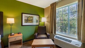 A room with a green wall, a framed picture, a brown leather chair, a wooden table, a nightstand with a lamp, and a window with curtains. - Extended Stay America Suites Clearwater Carillon Park (Clearwater)