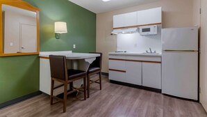 A compact kitchen with a dining table and chairs, a microwave, a refrigerator, and a wall-mounted lamp. - Extended Stay America Suites Clearwater Carillon Park (Clearwater)