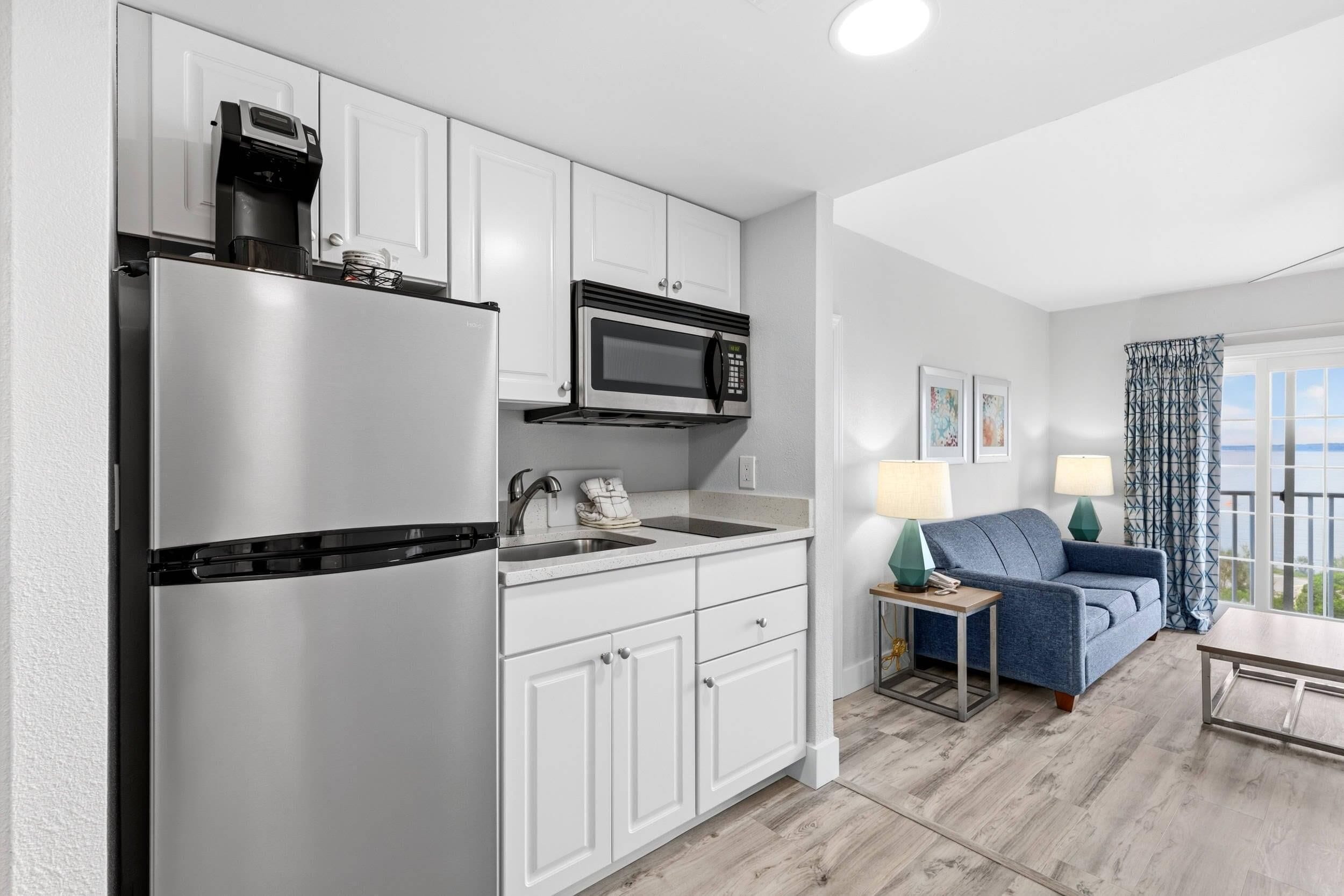 A modern kitchen with a refrigerator, microwave, and sink. There is a blue sofa and a table with lamps in the living area.
