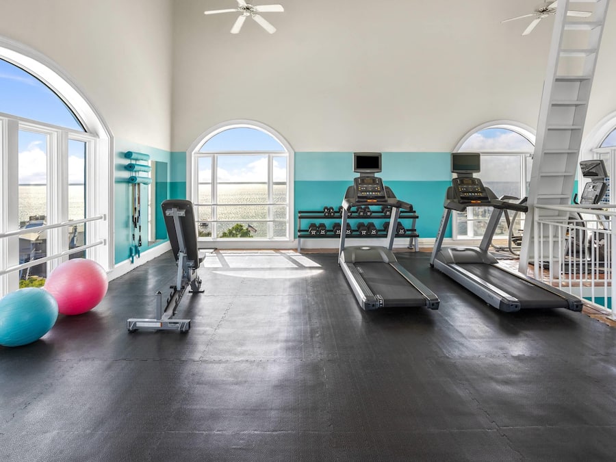 Academia com esteiras, halteres e bolas de exercício, grandes janelas com vista para o oceano.