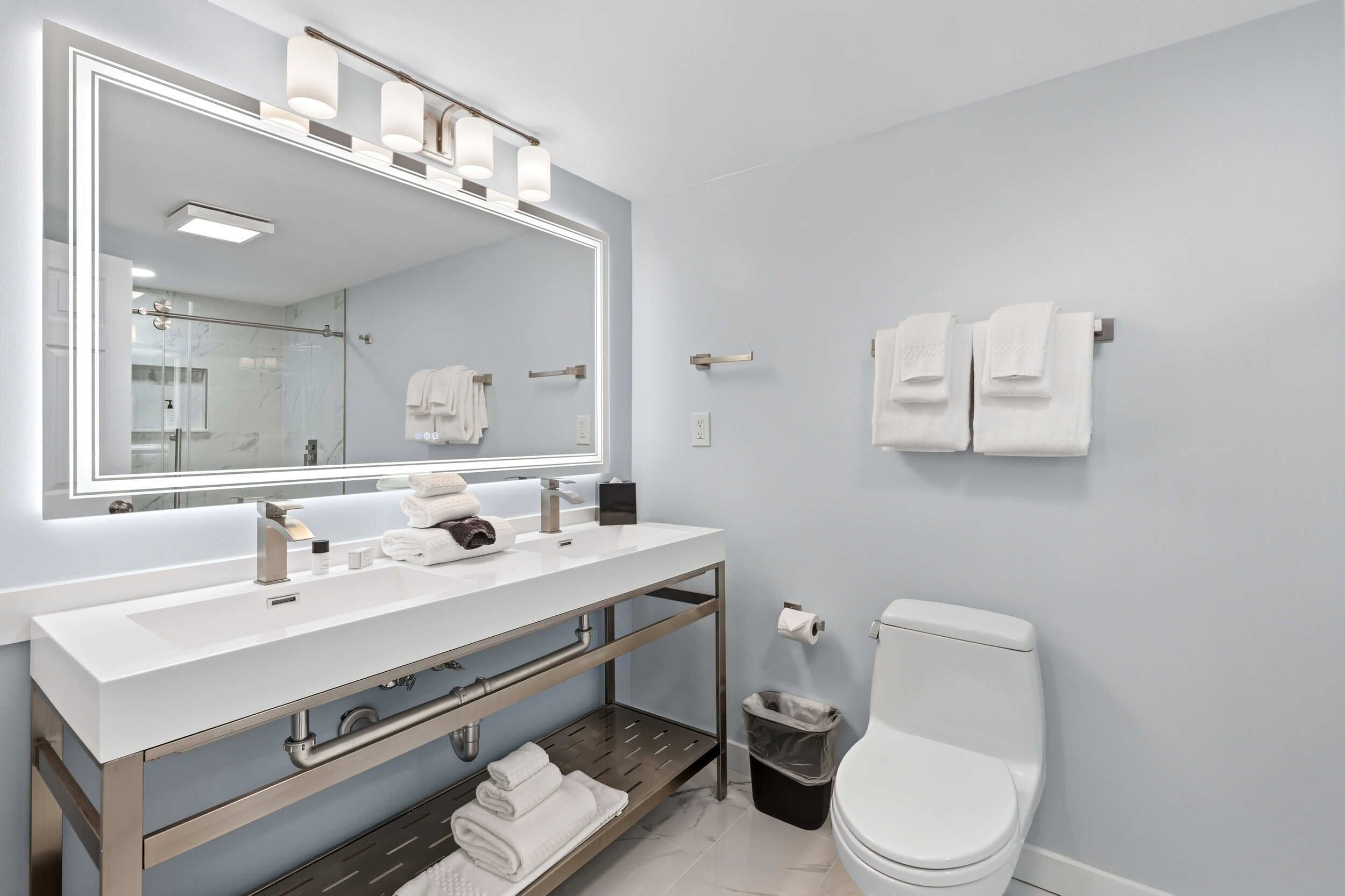Ein modernes Badezimmer mit großem Spiegel, zwei Waschbecken, Toilette und Mülleimer.