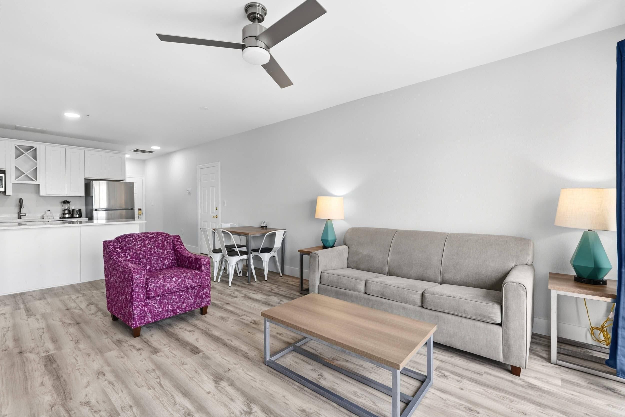 Un salon moderne avec un canapé gris, une table basse et un fauteuil violet. On y trouve également un coin repas avec une table et des chaises, et une cuisine à l’arrière-plan.