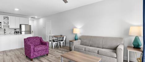 Un salon moderne avec un canapé gris, une table basse et un fauteuil violet. On y trouve également un coin repas avec une table et des chaises, et une cuisine à l’arrière-plan.