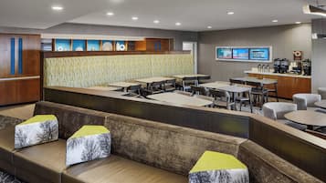 A modern hotel lobby with a seating area, a counter with a coffee machine, and a TV screen displaying content.