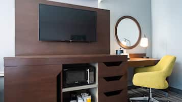 A hotel room with a wooden desk, a yellow chair, a flat-screen TV, a microwave, a coffee maker, and a lamp.