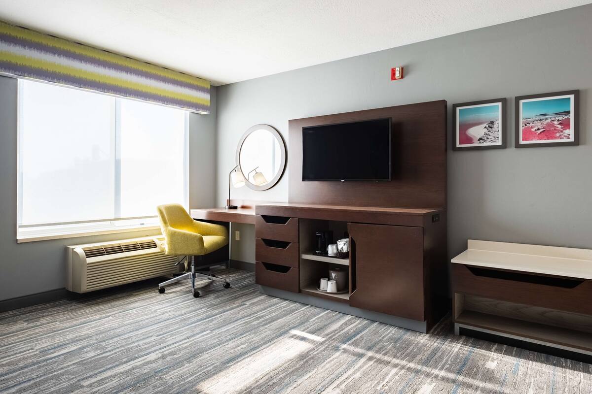 Ein Hotelzimmer mit großem Fenster, Schreibtisch mit Flachbildfernseher, Sessel, Heizung und zwei gerahmte Bilder an der Wand.