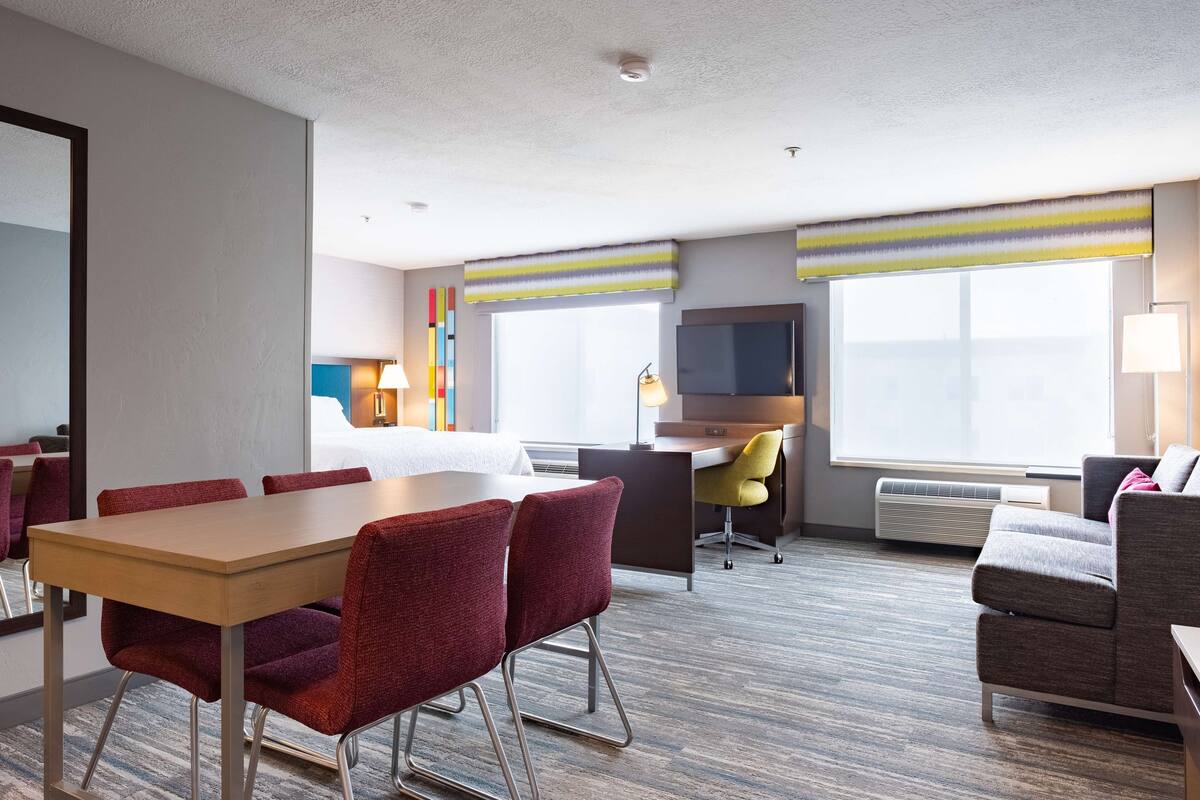 Ein Hotelzimmer mit Esstisch, roten Stühlen, einer Couch, einem Schreibtisch mit Stuhl, einem Fernseher und großen Fenstern mit gestreiften Vorhängen.