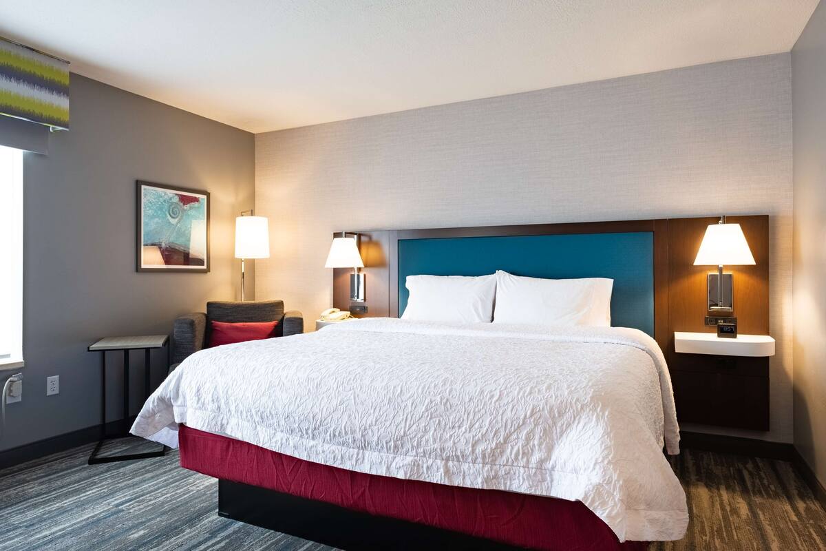 Ein Hotelzimmer mit einem großen Bett, zwei Nachttischlampen, einem kleinen Tisch, einem Sessel und einem Bild an der Wand.