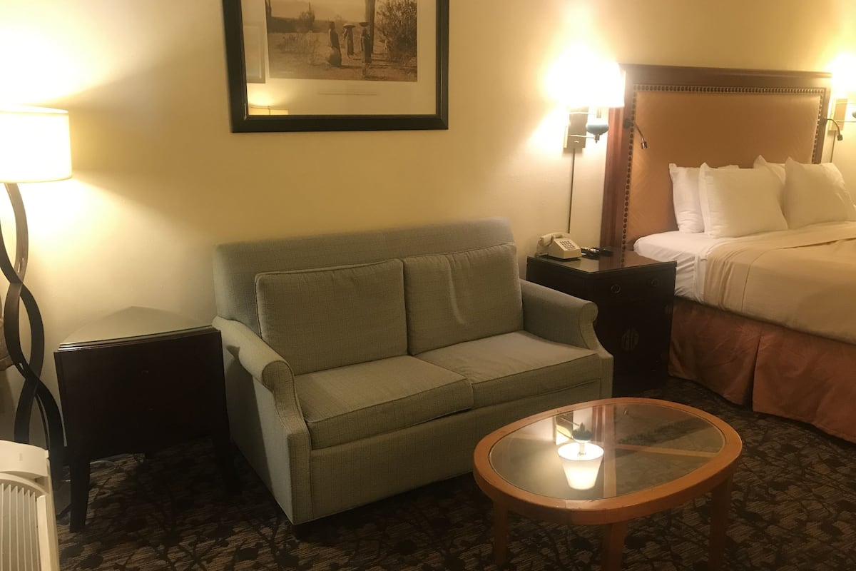 A hotel room with a green sofa, a glass coffee table, a bed with white linens, and a framed picture on the wall.