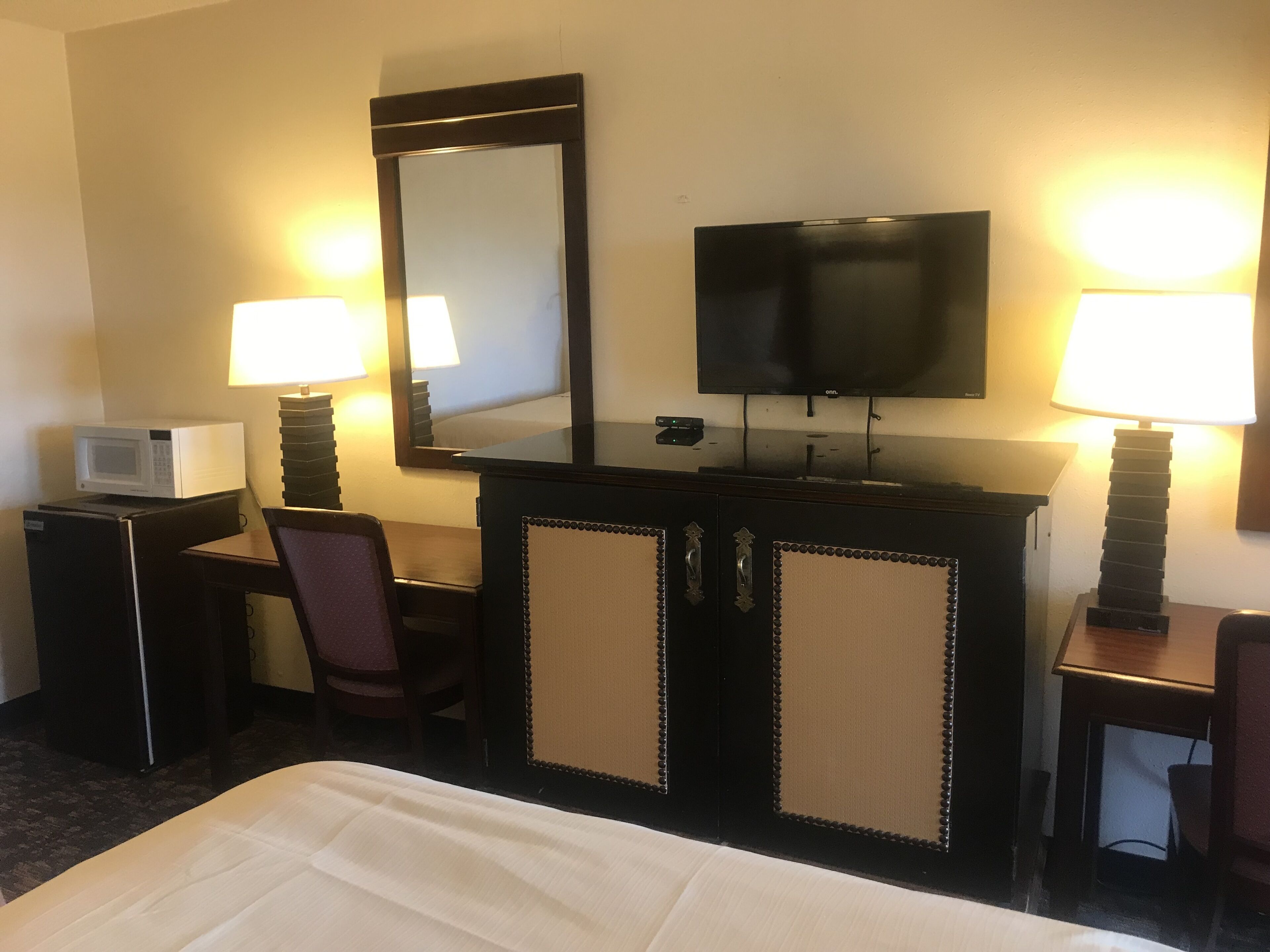 A hotel room with a bed, a desk with a television, a chair, a small table with a lamp, and a microwave.
