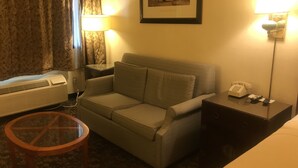 A hotel room with a sofa, a glass coffee table, a lamp, a framed picture on the wall, and a bedside table with a phone and remote control.