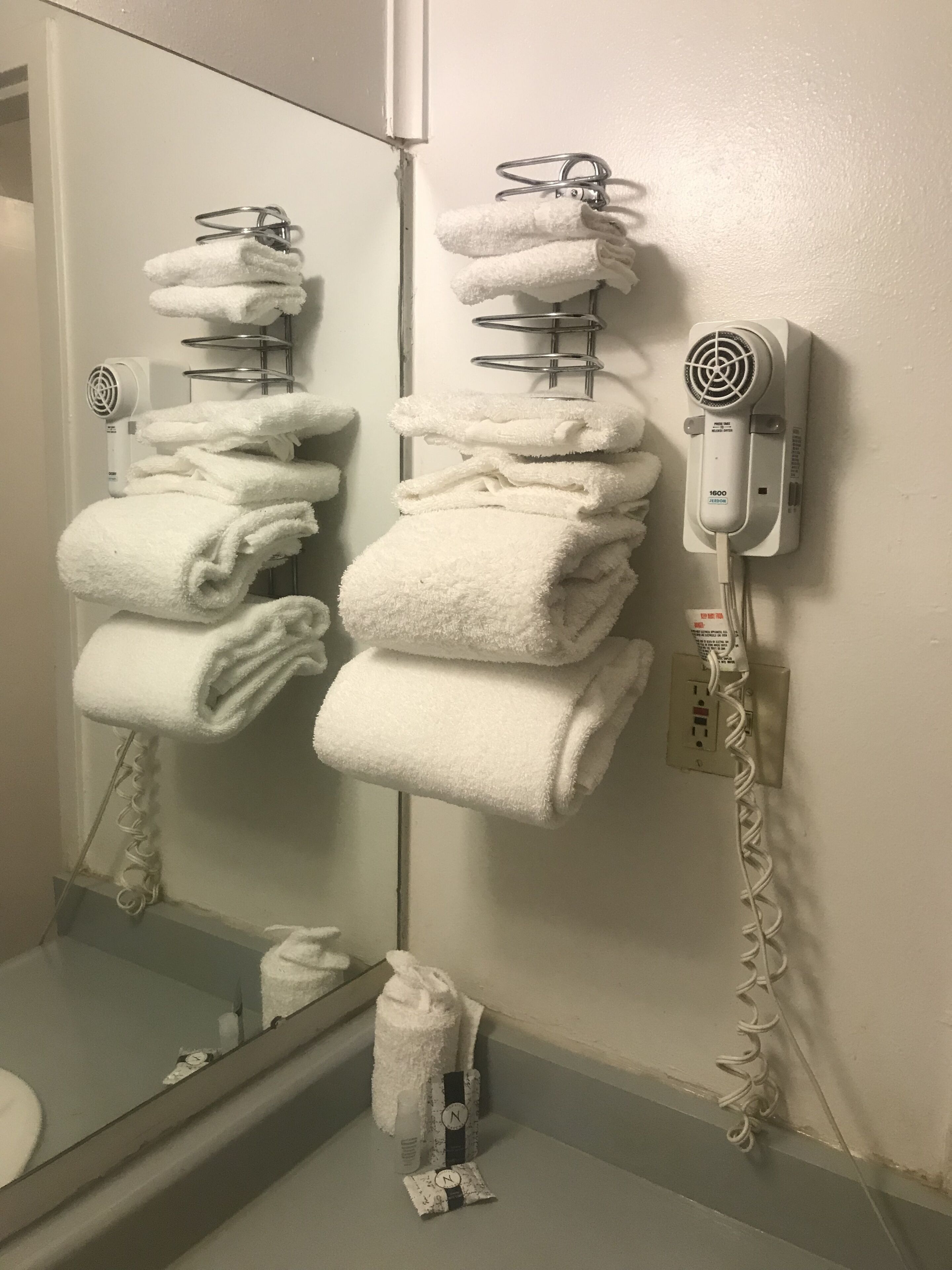 Un baño con un perchero con varias toallas dobladas, un secador de pelo en un soporte de pared y un espejo que refleja parte del cuarto.