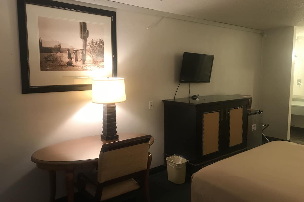 A hotel room with a bed, a desk with a lamp, a television on a cabinet, and a framed picture on the wall.