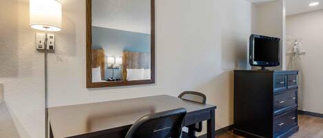 A hotel room with a desk, chair, TV, dresser, and a mirror.