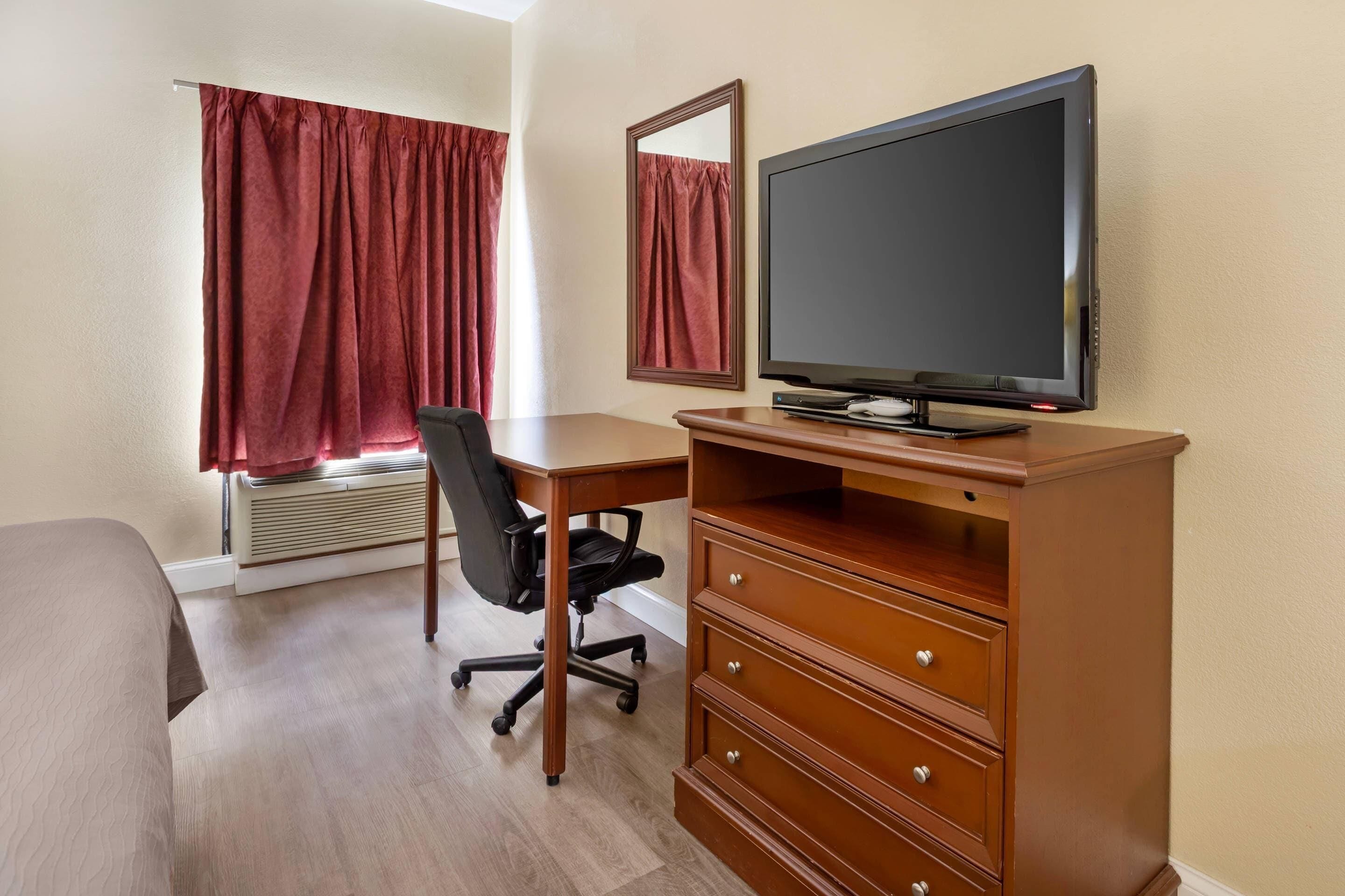 A hotel room with a wooden desk, a black office chair, a flat-screen TV, a mirror, and a large window with red curtains.