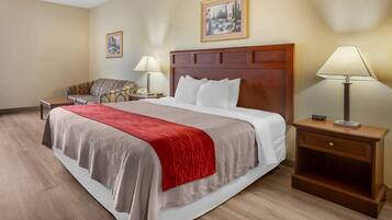 A hotel room with a large bed, a wooden headboard, a red throw blanket, a nightstand with a lamp, a chair, and a framed picture on the wall.