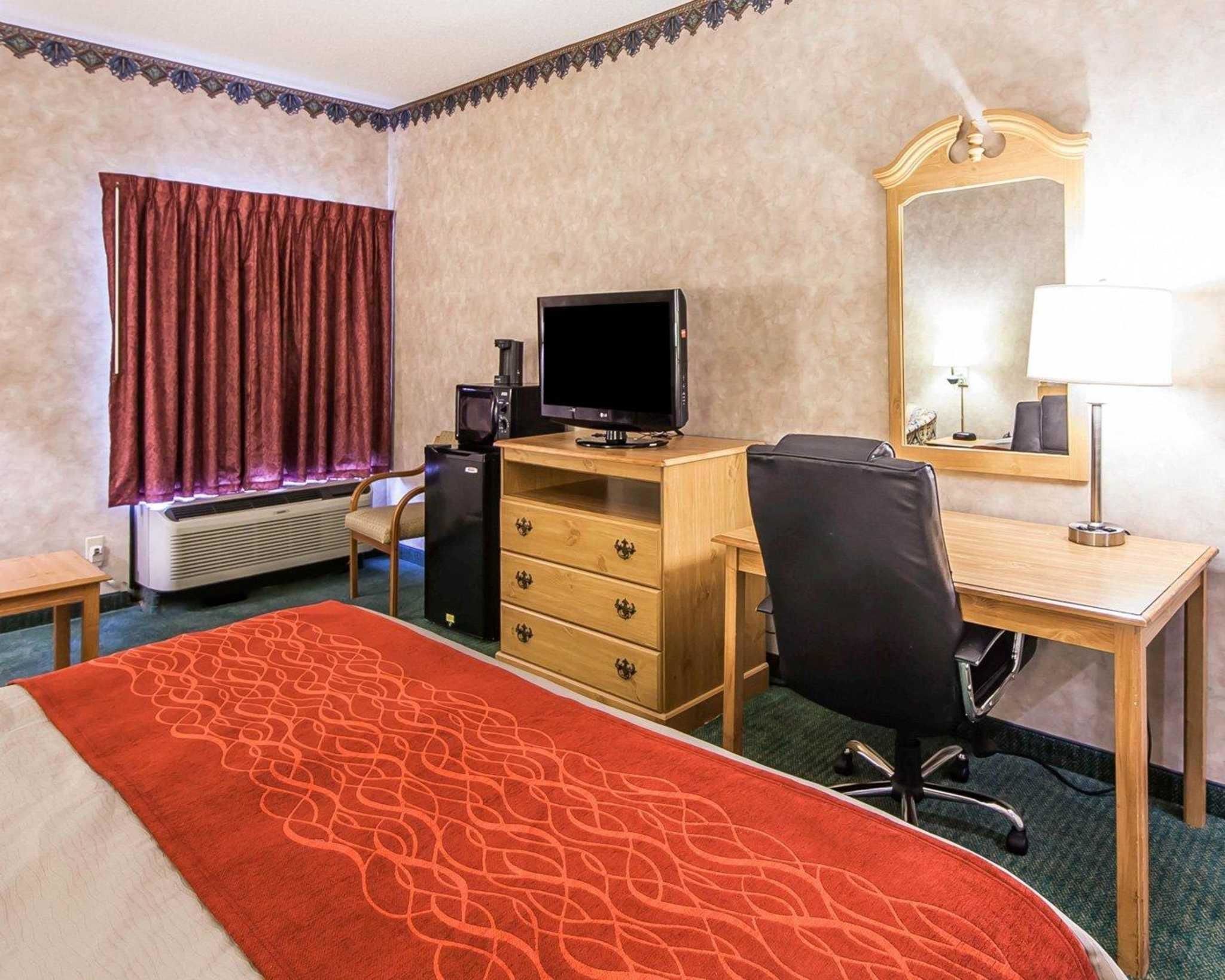 A hotel room with a bed, a desk with a computer, a chair, a TV, a mirror, and a window with curtains.