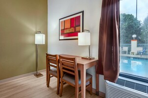 Habitación de hotel con un escritorio de madera, dos sillas, un cuadro abstracto enmarcado, dos lámparas de pared y vistas a una piscina a través de una ventana con cortinas marrones.
