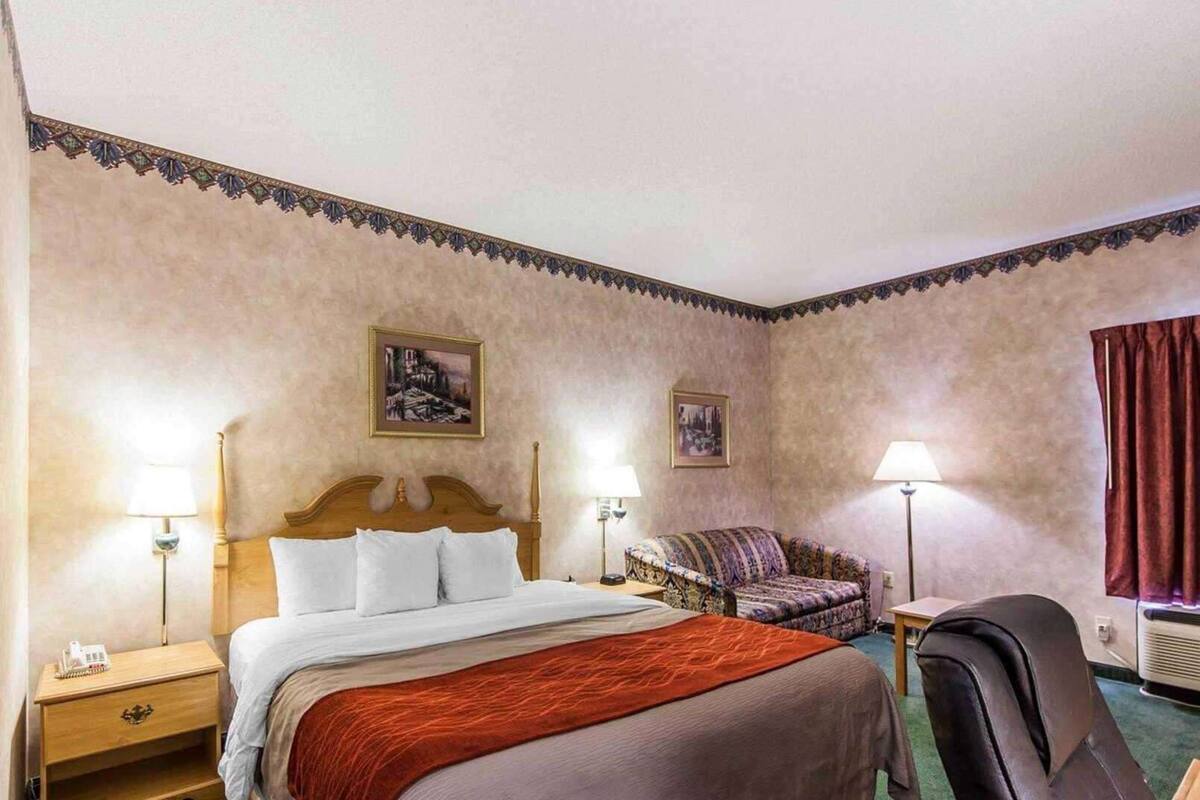 A hotel room with a bed, a sofa, a chair, two lamps, and two paintings on the walls.