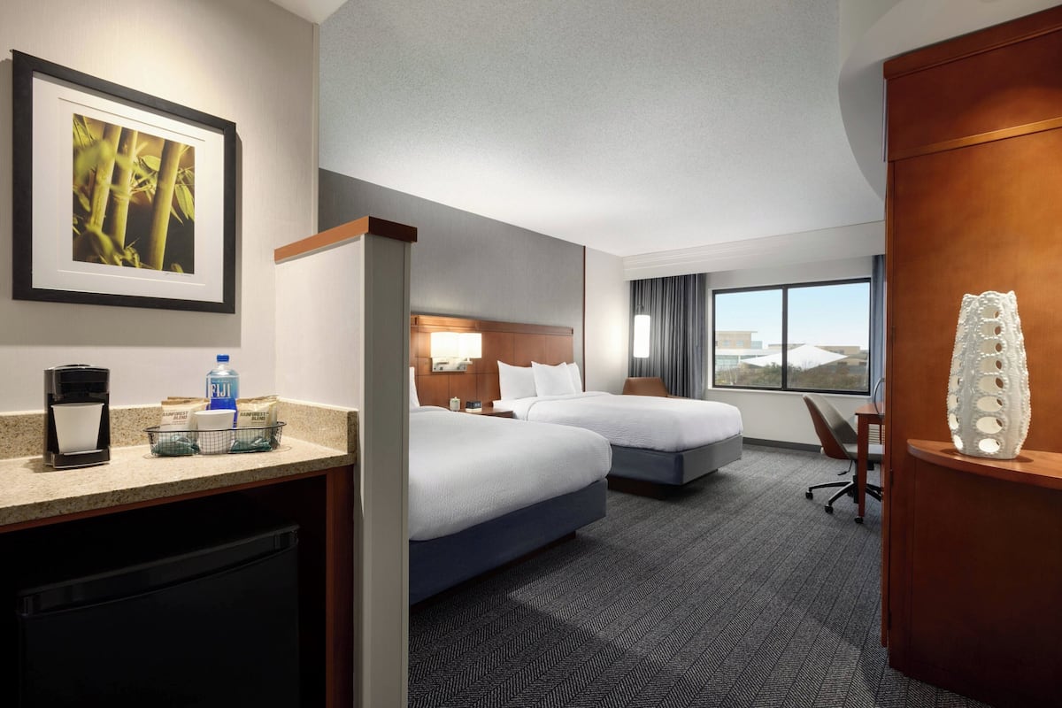 A hotel room with two beds, a desk with a coffee maker, a trash bin, a water bottle, a framed picture, and a large window with a view.