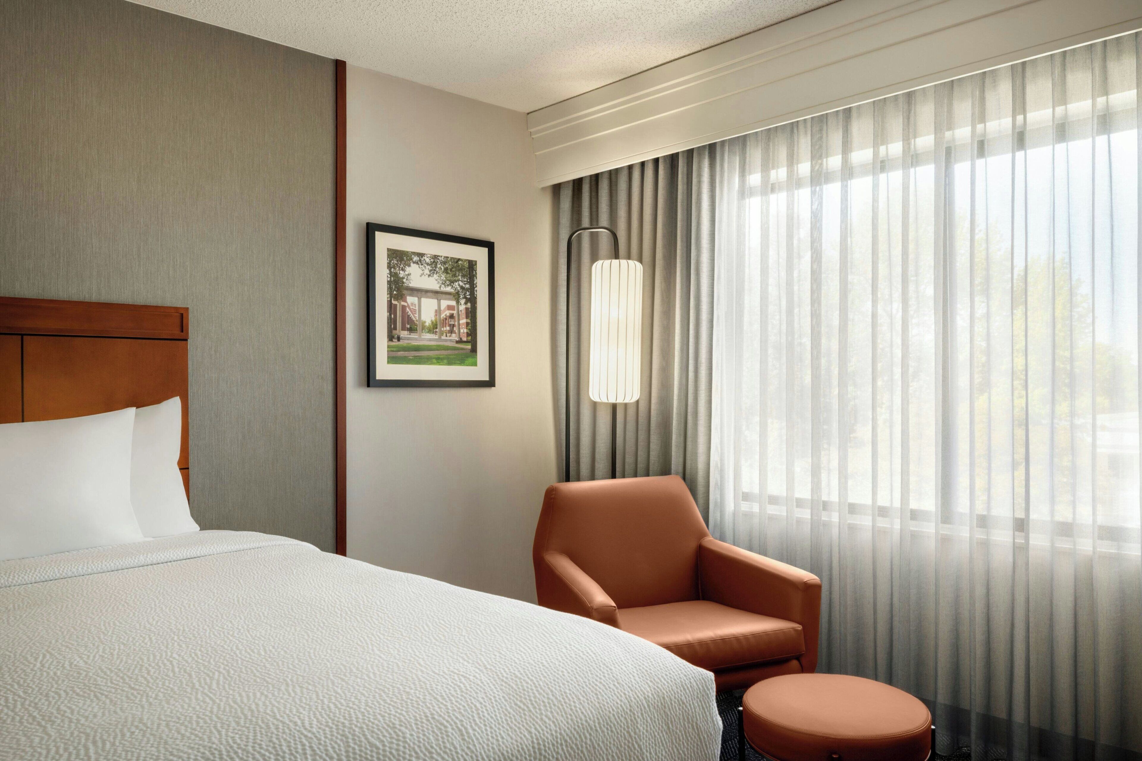A hotel room with a large bed, a brown armchair, a small round table, a framed picture on the wall, and a window with sheer curtains.
