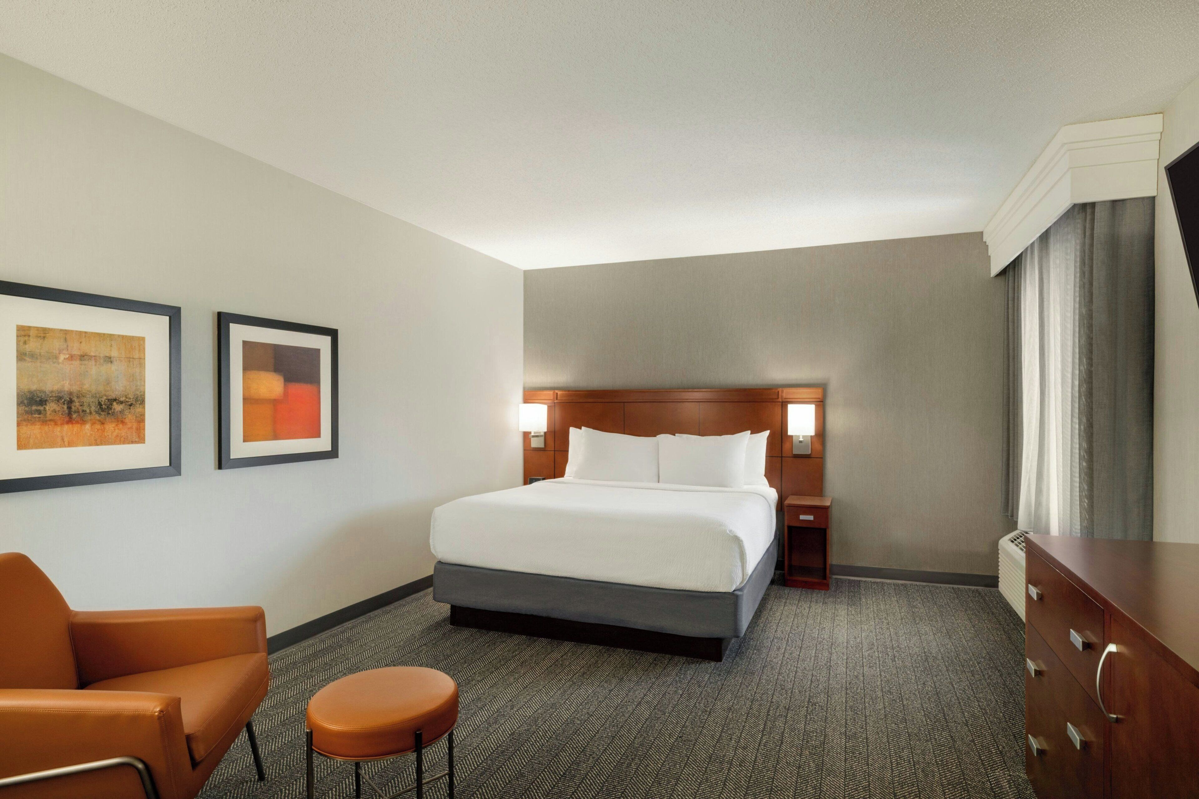 Habitación de hotel con una cama, una silla, una mesita y dos cuadros en la pared.