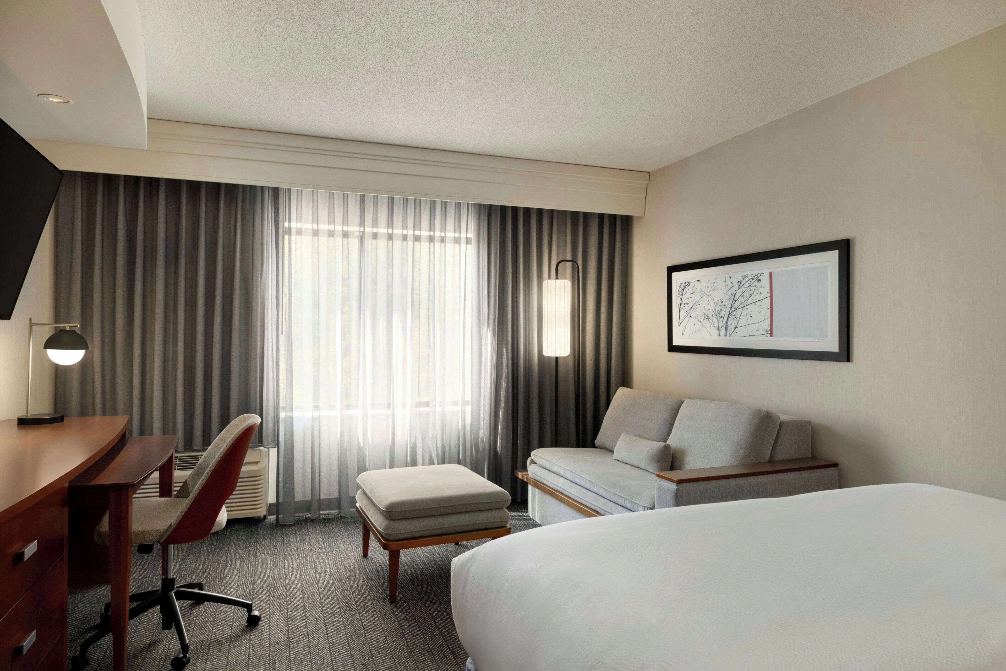 A hotel room with a bed, a desk, a chair, a sofa, and a large window with curtains.