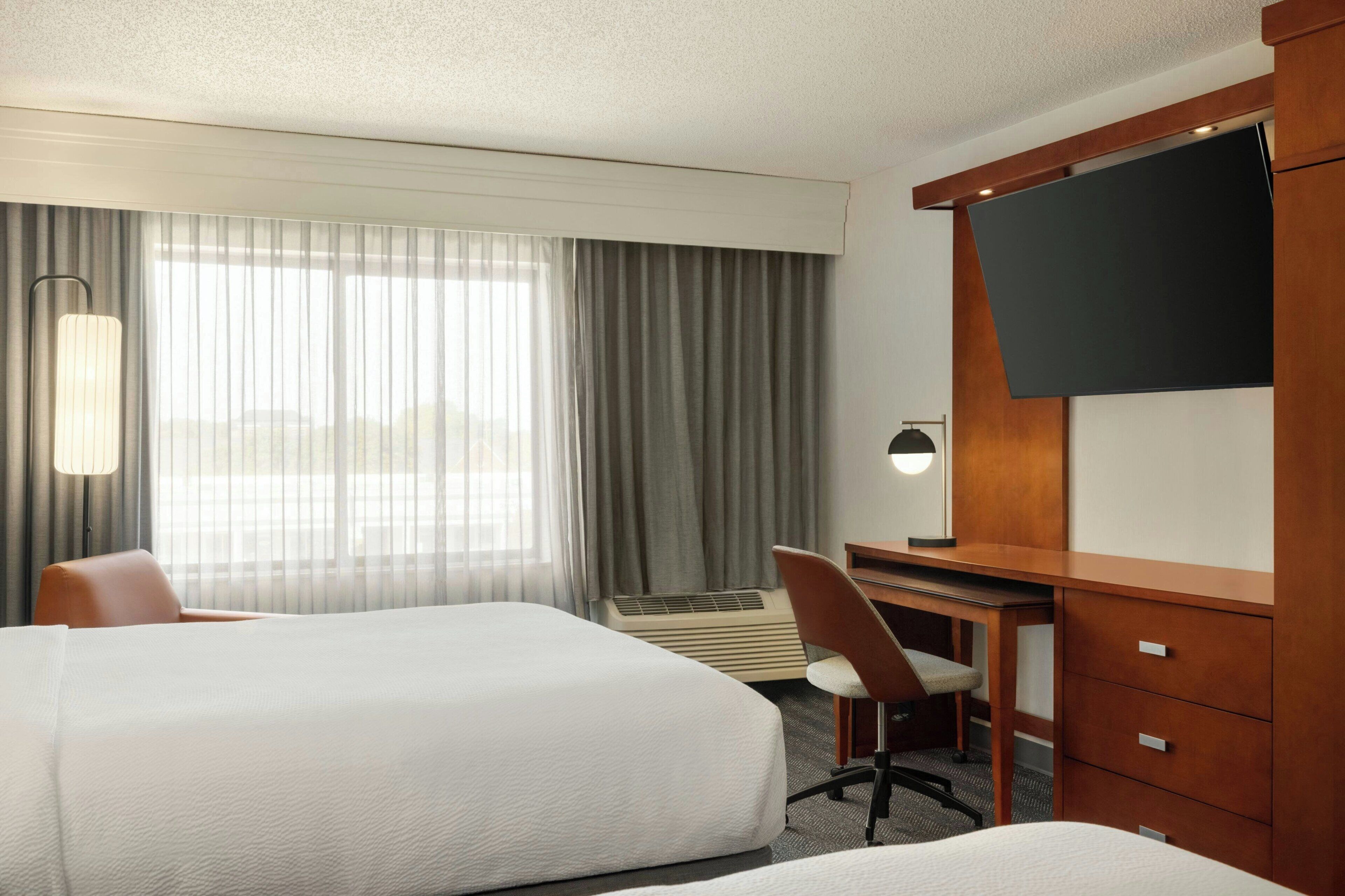 Ein Hotelzimmer mit zwei Betten, einem Schreibtisch, einem Sessel, einem Fernseher und einem großen Fenster mit Vorhängen.