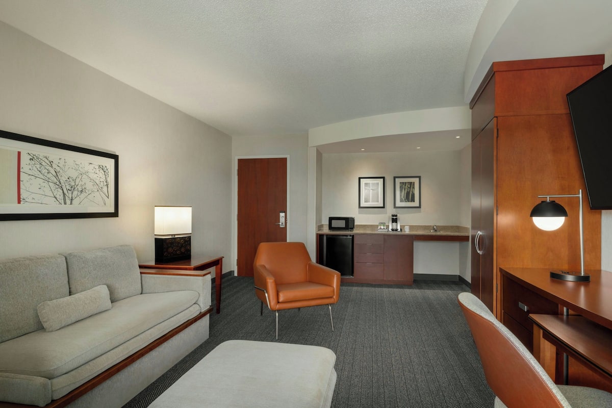 A hotel room with a sofa, armchair, ottoman, kitchenette, and a flat-screen TV.