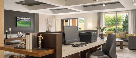A modern office with a desk, computer, and ergonomic chair. There is a television on the wall and a window with a view of greenery.