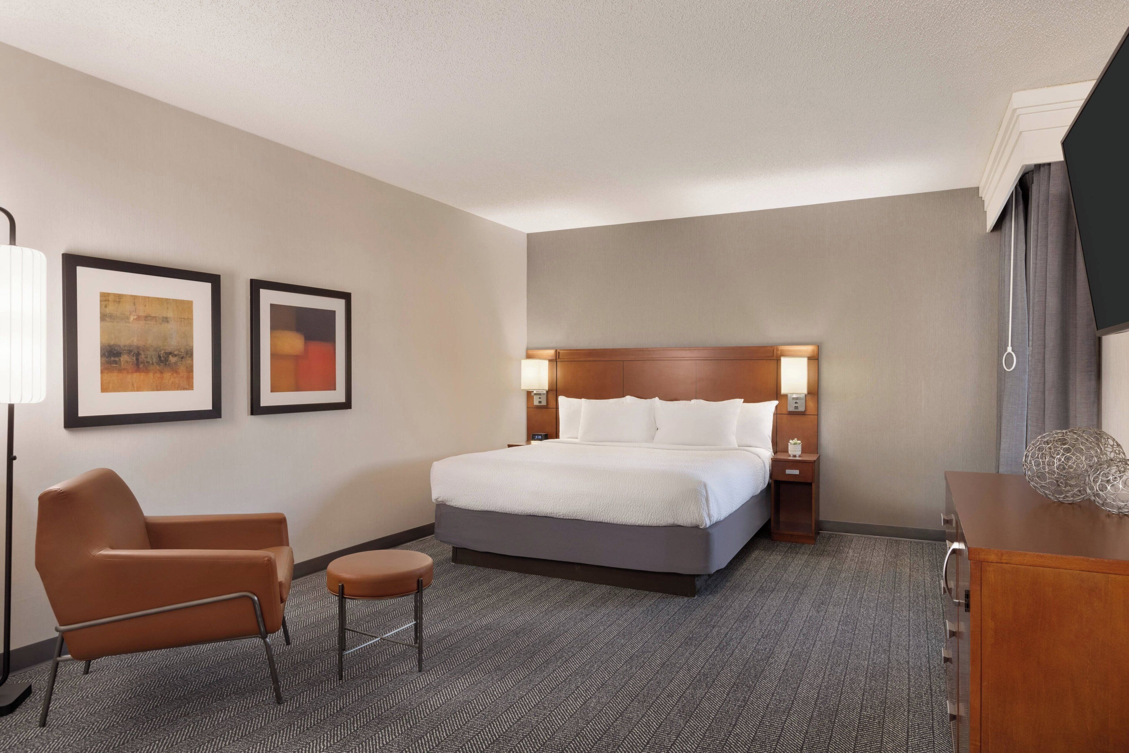 A hotel room with a large bed, a chair, a small table, and two framed pictures on the wall.