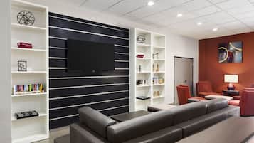 A modern living room with a large flat-screen TV mounted on a black wall, surrounded by white shelving units with books and decorative items. There is a grey sofa and a small table with a lamp.