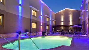 A swimming pool with a sunken seating area, surrounded by lounge chairs and umbrellas, with a building in the background. - Country Inn & Suites by Radisson, San Antonio Lackland AFB - SeaWorld Area (San Antonio)