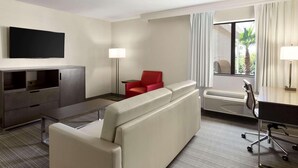 A hotel room with a sofa, a red chair, a desk, a flat-screen TV, a lamp, and a window with curtains. - Country Inn & Suites by Radisson, San Antonio Lackland AFB - SeaWorld Area (San Antonio)