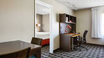 Quarto de hotel com cama, escrivaninha, cadeira, luminária e janela com cortinas.