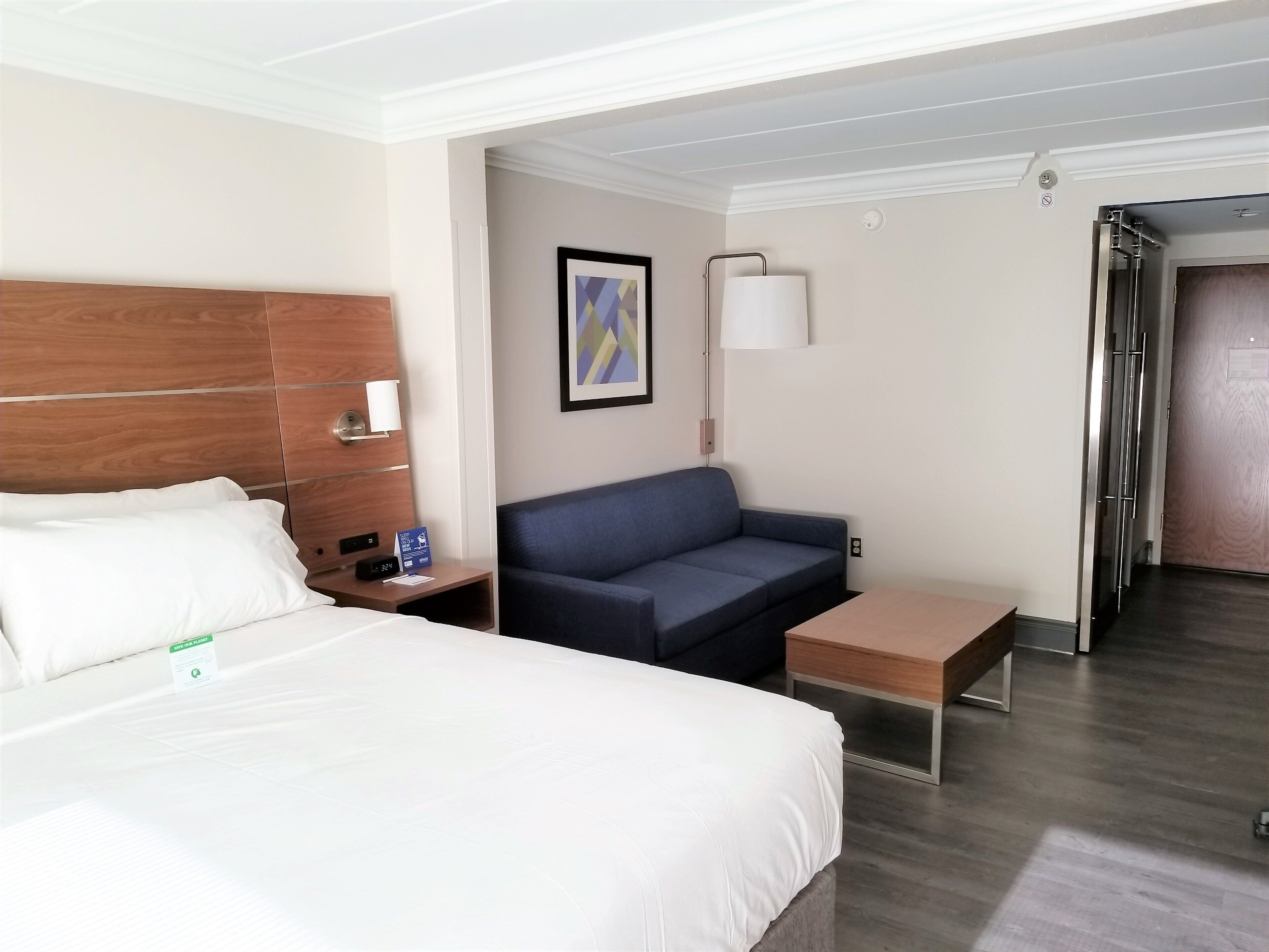 Habitación de hotel con cama, sofá, una mesita, una lámpara de pared y una puerta que da a otra habitación.