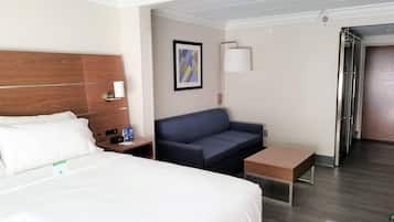 Habitación de hotel con cama, sofá, una mesita, una lámpara de pared y una puerta que da a otra habitación.