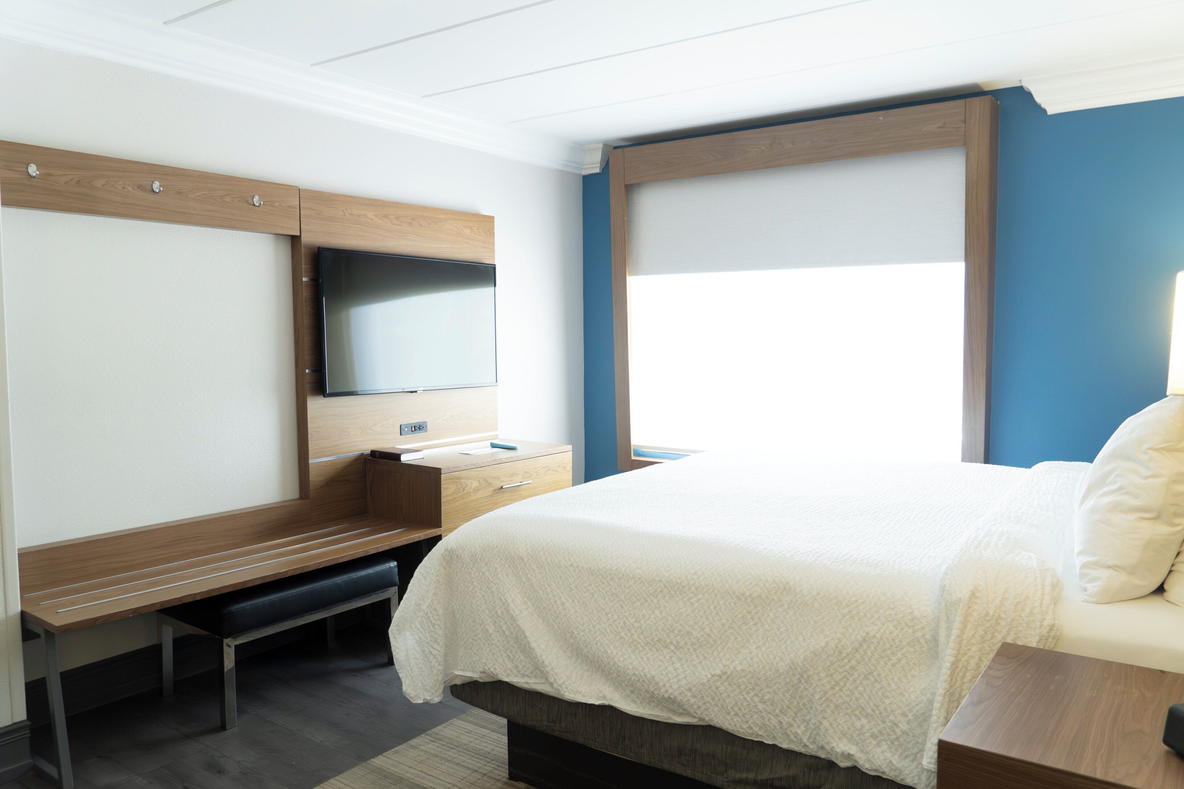 A hotel room with a bed, a desk, a flat-screen TV, a window, and a wooden headboard.
