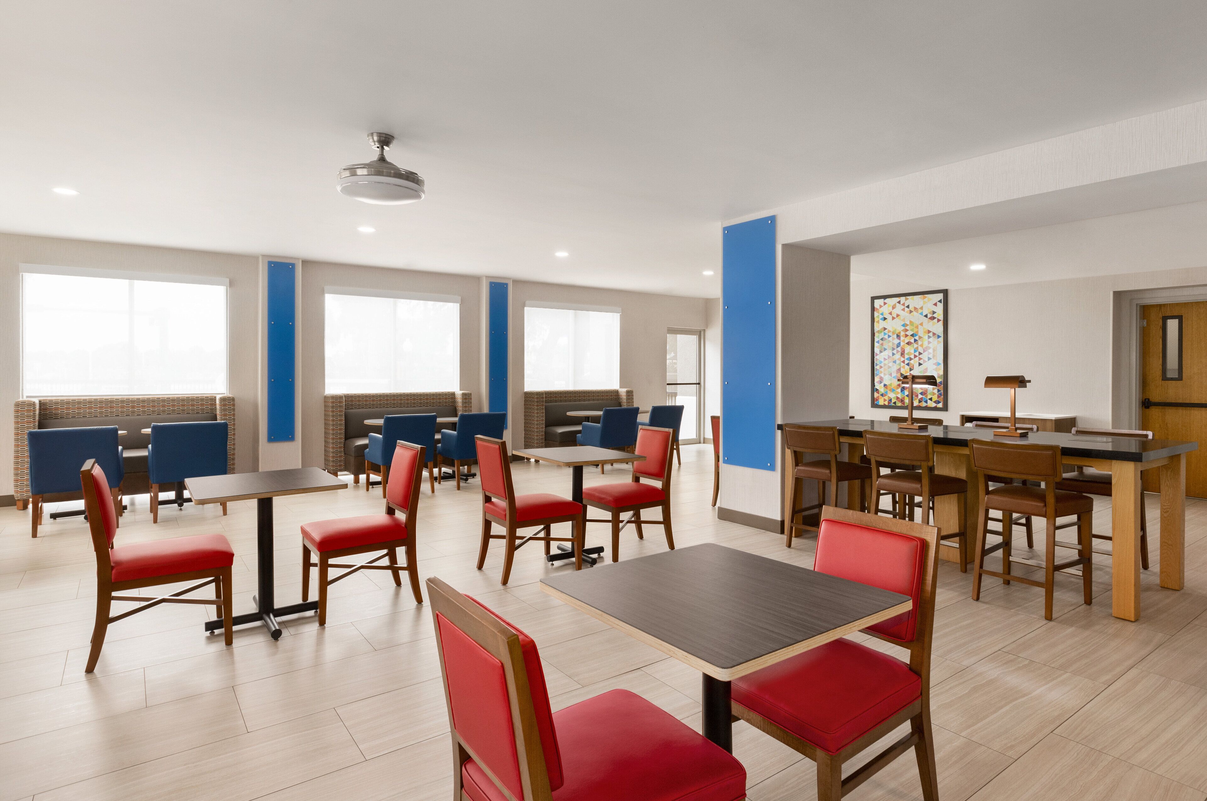 Un espace repas moderne doté de plusieurs tables et chaises, de grandes fenêtres et d’un ventilateur de plafond.