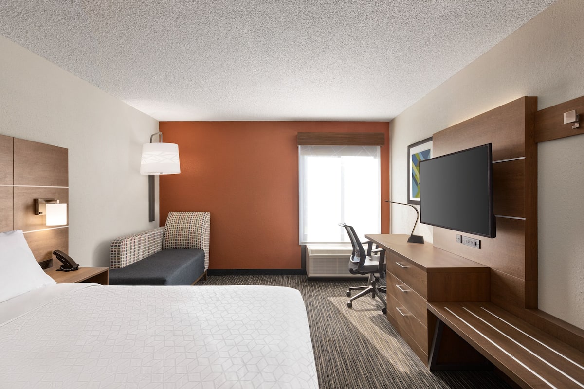 A hotel room with a large bed, a desk with a flat-screen TV, a chair, and a window with blinds.