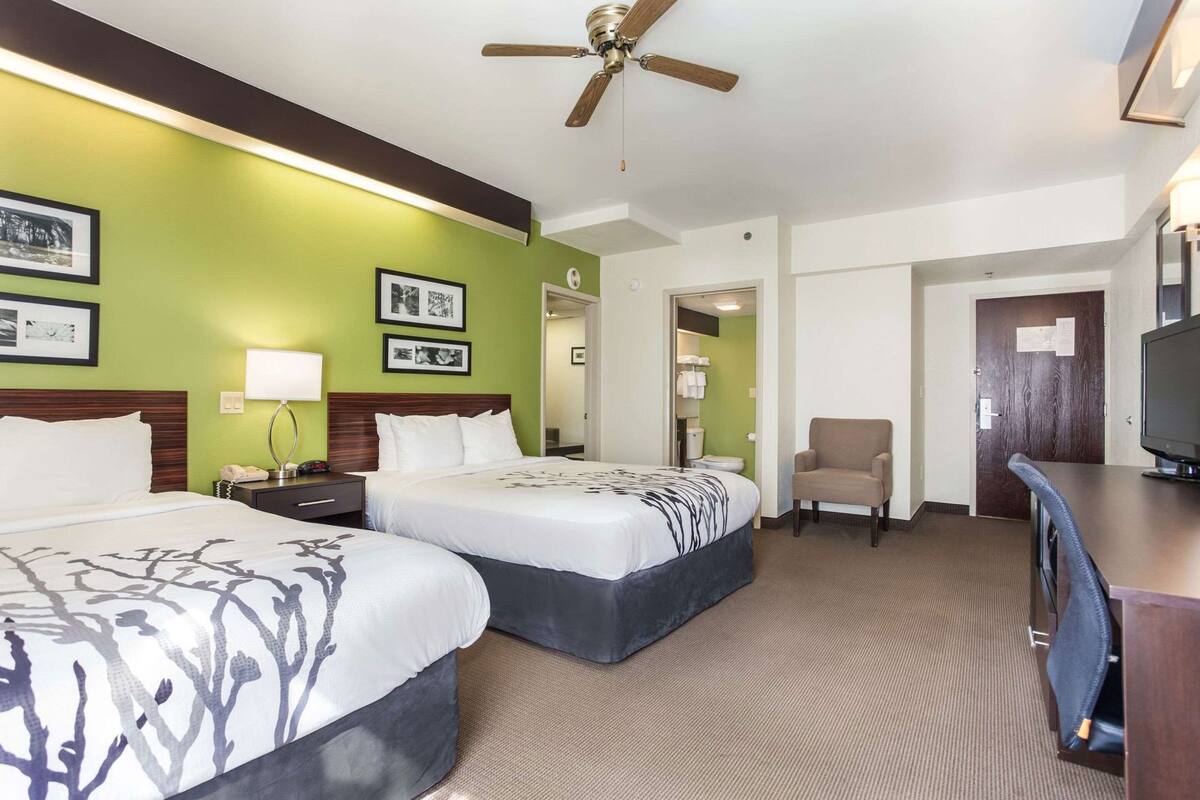 A hotel room with two beds, a desk, a chair, a television, and a ceiling fan.