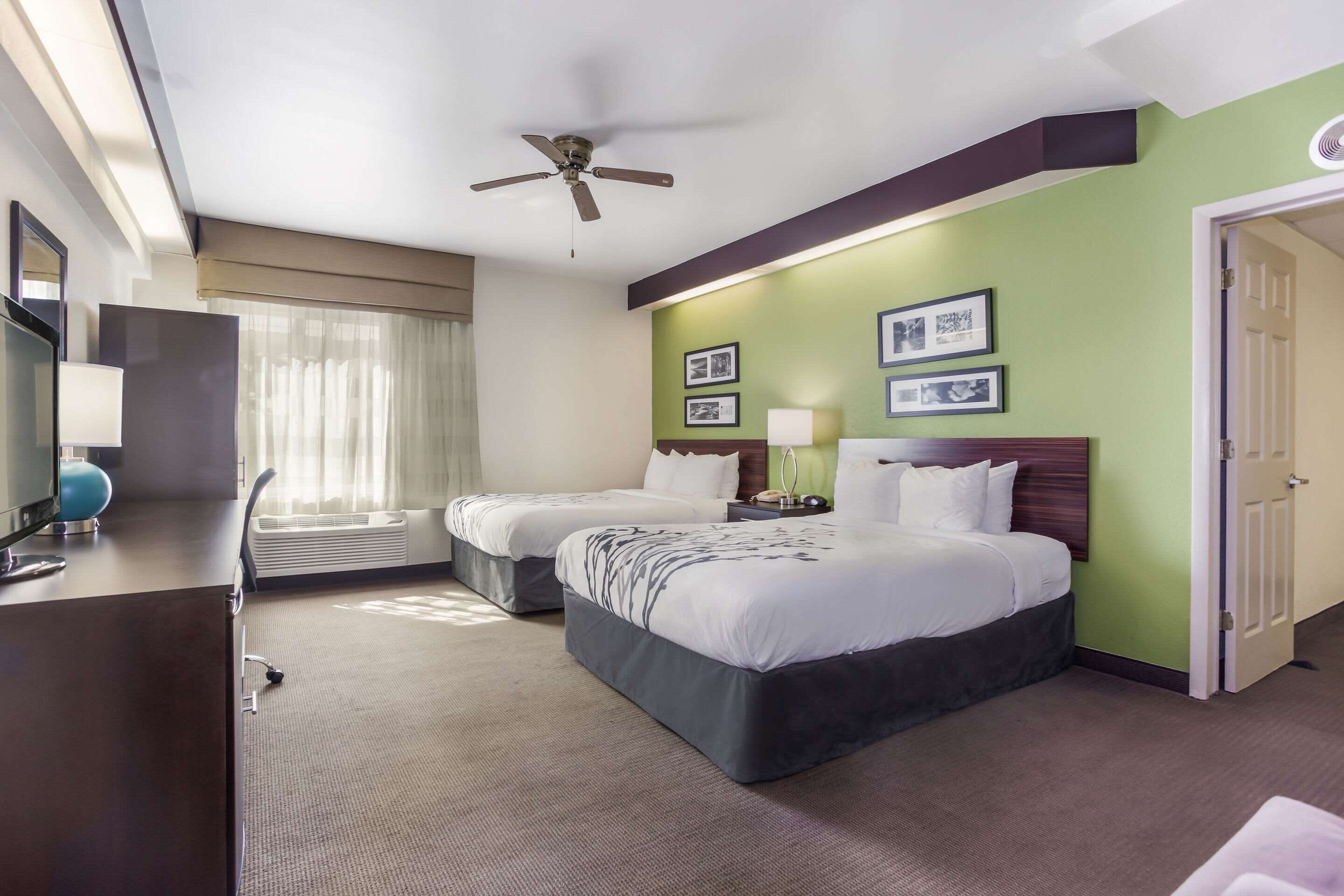 A hotel room with two beds, a desk with a lamp, a ceiling fan, and framed pictures on the wall.