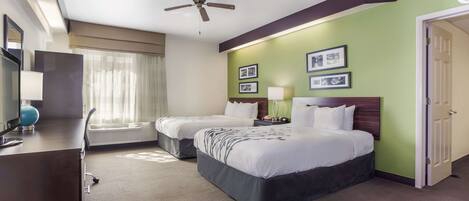 A hotel room with two beds, a desk with a lamp, a ceiling fan, and framed pictures on the wall.