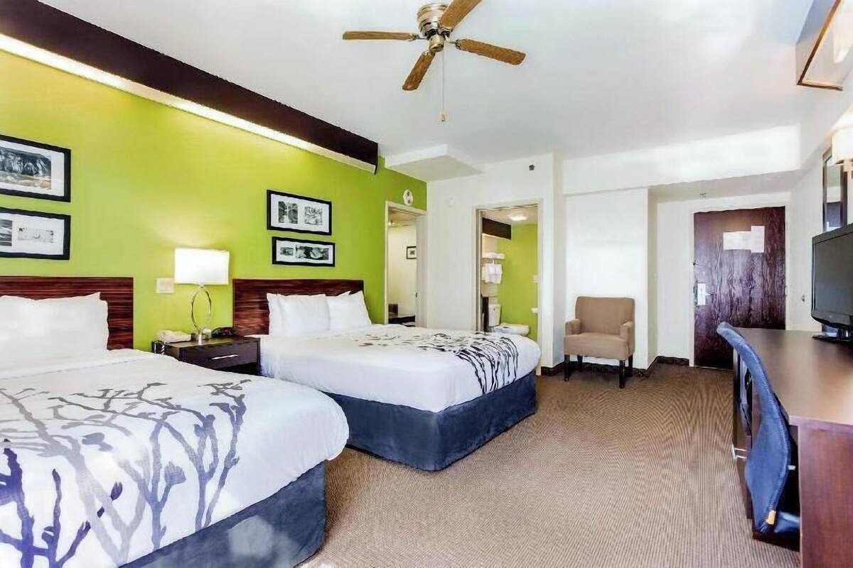 A hotel room with two beds, a desk, a chair, a television, and a ceiling fan.