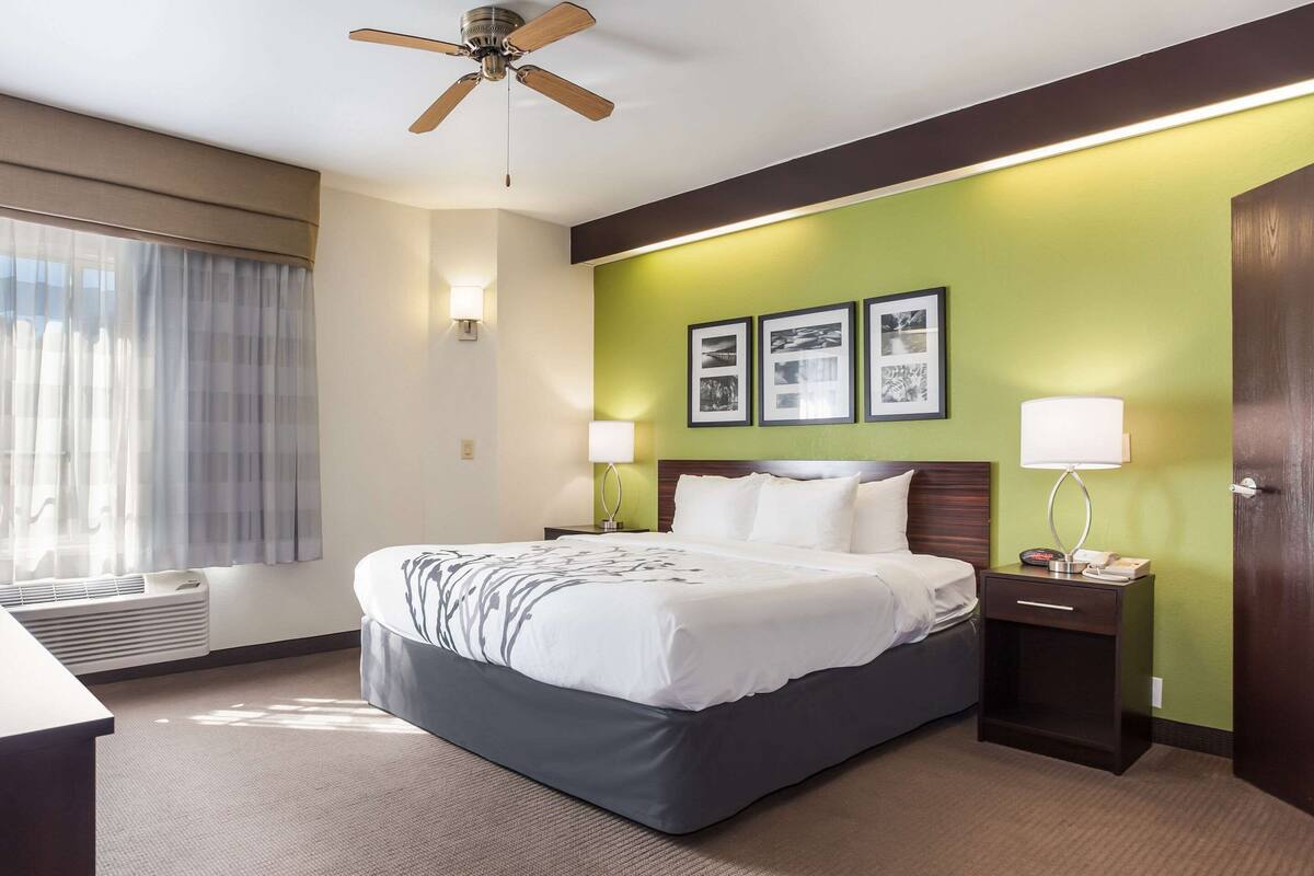 A hotel room with a bed, a nightstand, a lamp, a ceiling fan, and a window with curtains.