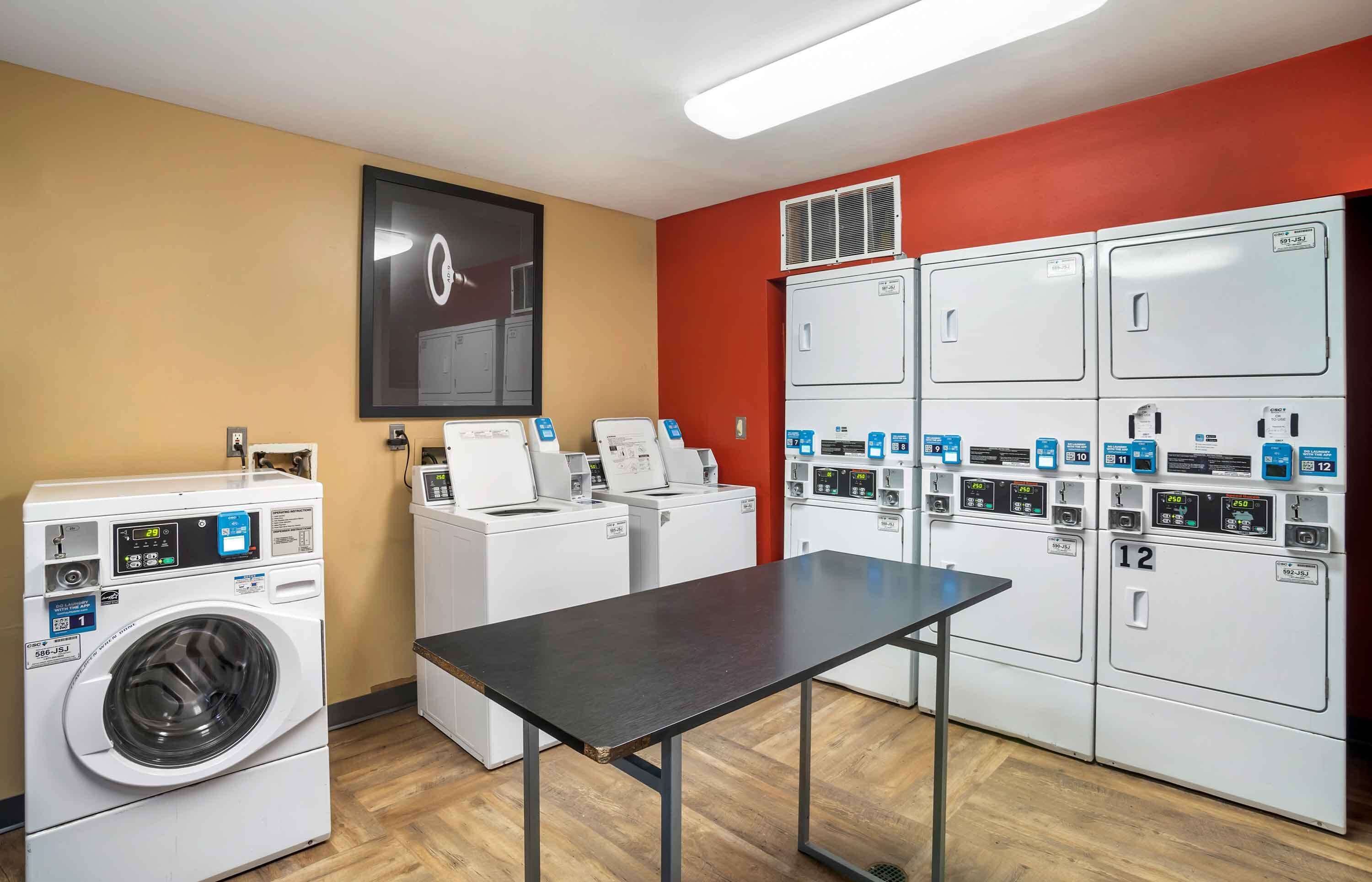 A laundry room with multiple washing machines and dryers, a table, and a wall-mounted light fixture.