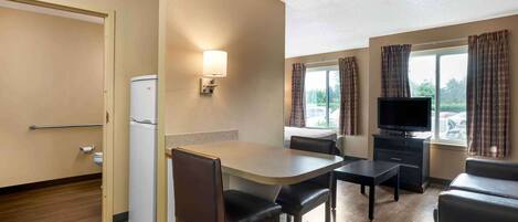 Et hotelværelse med tekøkken, spiseplads med bord og stole, sofa, fjernsyn og et vindue med gardiner.